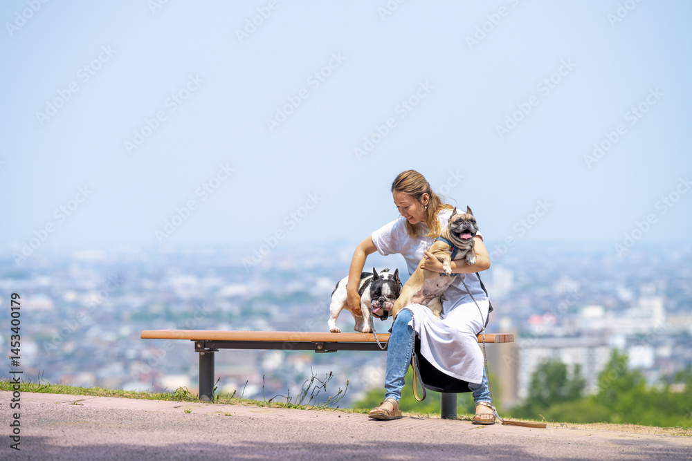 On a summer morning in a park with blooming hydrangeas, a Japanese woman in her 30s walks two French Bulldogs. They sit on a hilltop bench, overlooking the city, with flowers and greenery all around.