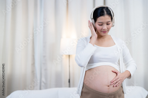 Asian pregnant woman wearing headphones smiling softly eyes closed gently cradling belly standing in white top beige pants feeling calm connection joy love bonding with unborn baby during pregnancy