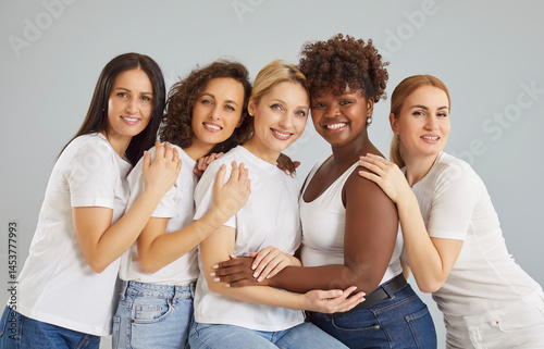 Isolated shot of happy mixed race women stand against grey wall closely to each other and embracing, looking directly at camera, have toothy smile, clean skin. Woman beauty concept