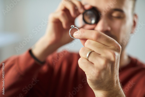 Closeup portrait of young man jeweler looking through magnifying glass examining gold ring evaluating jewelry with diamond. Jewellery appraiser working with jewel. Selective focus.