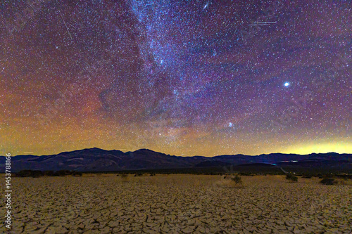 The Milky Way arches over cracked desert ground and mountains in Death Valley National Park under a clear night sky.