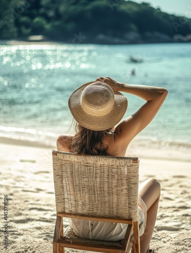 A woman sitting in a wicker chair with her legs crossed, wearing a straw hat, sipping a drink and looking out at the ocean.
