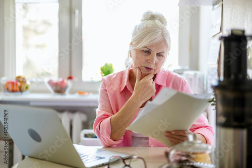 Senior woman working with laptop and documents at kitchen table at her home
