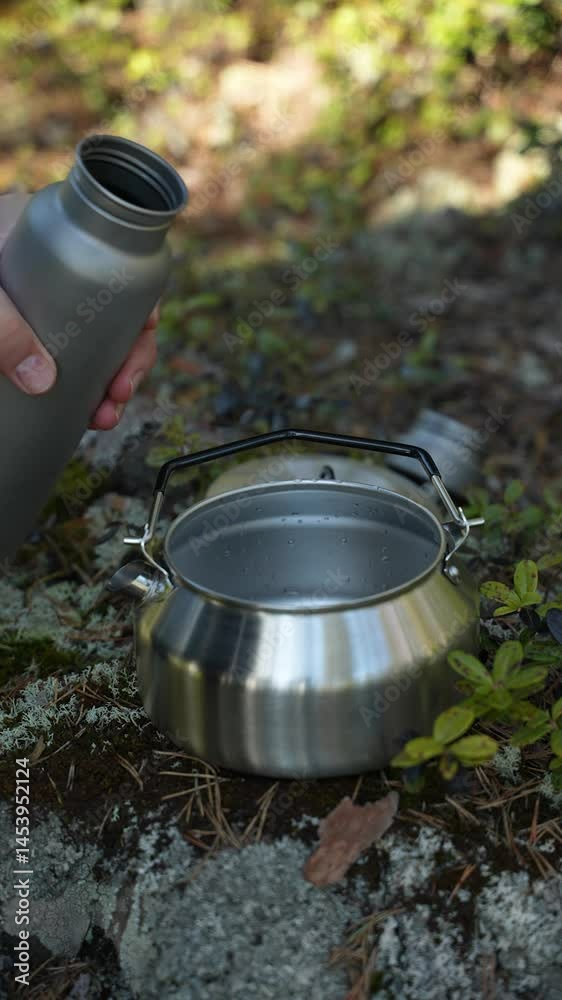 A tourist is pouring water from a titanium flask into a camping kettle. Making tea while hiking in the forest. Gear for tourism and hiking. Close-up shot. A tourist is pouring water from a titanium flask into a camping kettle. Making tea while hiking in the forest. Gear for tourism and hiking. Close-up shot.