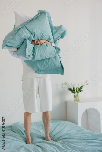 A person is currently standing on a bed while holding a fluffy pillow high above their head, creating a playful atmosphere in the room