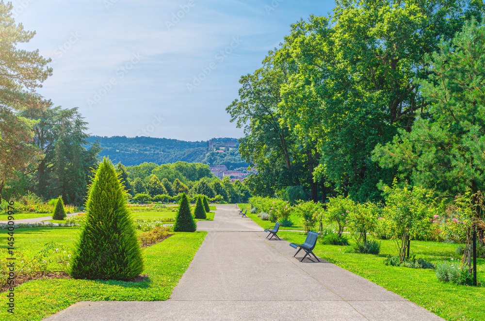 Parc des Glacis park with green trees and bushes, grass lawn and benches on path in Besançon city in sunny summer day with blue sky background, Bourgogne-Franche-Comte region, France