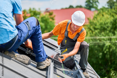 Worker building photovoltaic solar panel system on roof of house. Engineer in helmet looking on evenness of solar modules during installing outdoors. Concept of alternative and renewable energy.