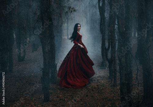 Art photo real person Gothic girl walking running in dark foggy forest trees, red long medieval old style long dress flies in wind, train fluttering in motion. Fantasy Woman Black hair beautiful face