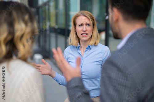 Fotografie Upset young woman in business attire raising hands in frustration while having a heated discussion with colleagues in a modern business district