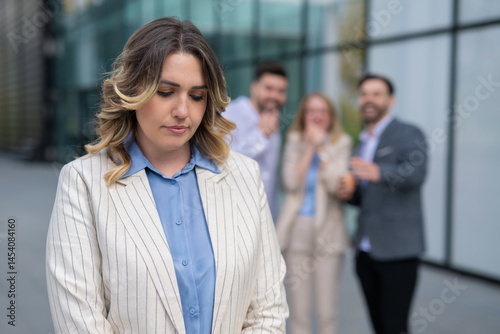 Sad businesswoman facing bullying at work while coworkers laugh behind her