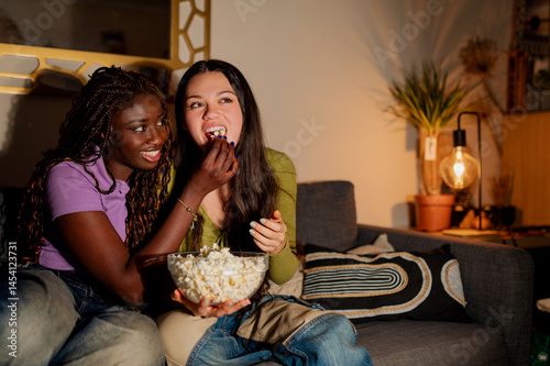 Two women are sitting on a sofa, sharing a bowl of popcorn and watching a movie in their living room