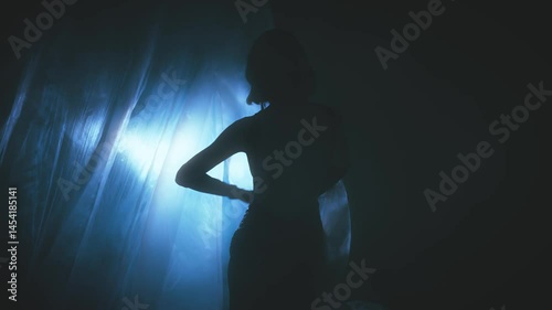 dark silhouette woman walking to neon blue light in night room full smoke fog silk curtains fly rain lightning wind blowing slow motion. Gothic Girl fashion model in sexy black dress. back rear view