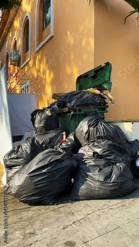 Pile of black garbage bags overflowing from a green dumpster on a city sidewalk under daylight.