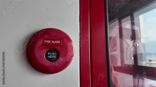 Slow motion footage of Red fire alarm on a wall with a PUSH HERE button in the center. It’s placed next to a glass cabinet containing fire safety equipment.