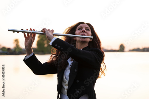 beautiful middle-aged woman is playing the transverse flute dressed in an elegant tailcoat at sunset on a small pier