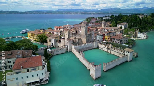 Drone approach to Scaliger Castle in Sirmione, Italy. Historic walls, colorful houses, and turquoise waters of Lake Garda captured on a sunny spring day.