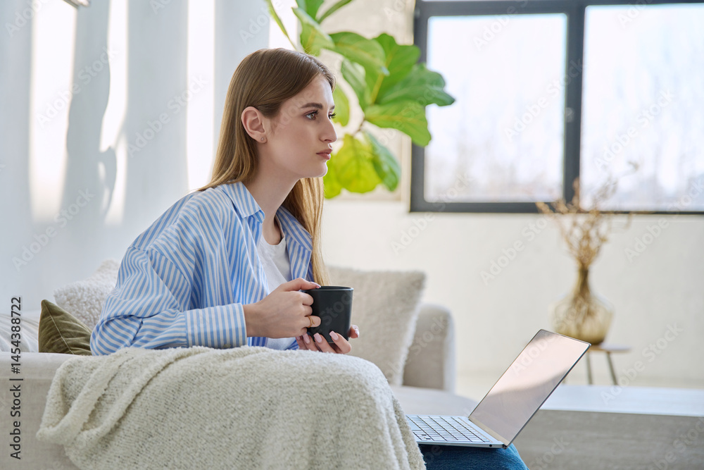 Portrait of young beautiful smiling blonde woman with cup of drink sitting on sofa at home