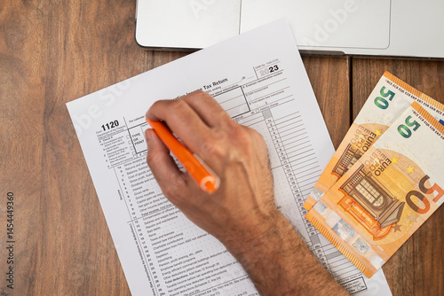 Business owner carefully fills out 1120 tax form on wooden table using laptop and referencing euro bills to ensure all foreign transactions properly recorded