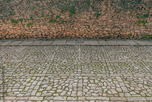 Fototapeta Naklejka Na Ścianę i Meble -  Close-up view of an old cobblestone street leading to a weathered stone wall with small green plants growing between the cracks.