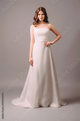 Sleeveless maxi wedding dress in white with pleated bodice, strapless neckline. Full-length studio portrait of young beautiful brunette bride. Bridal outfit in classic style, trendy wavy hairdo photo