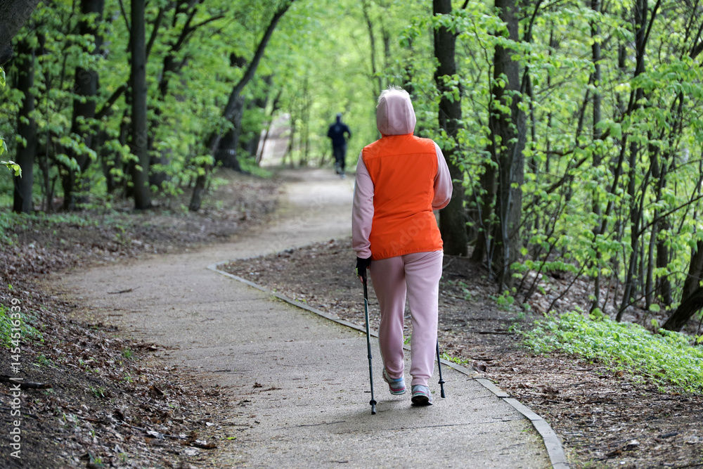 Nordic walking at spring weather, woman with sticks wearing sportswear walk in park on green trees background