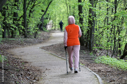 Obraz na płótnie Nordic walking at spring weather, woman with sticks wearing sportswear walk in park on green trees background w salonie