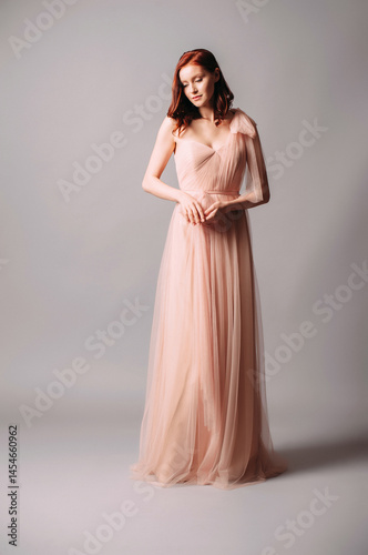 Elegant bridesmaid’s two-layer maxi gown. Gorgeous young sexy lady with curly hairstyle. Ivory evening pleated dress with bow detail. Seductive ginger woman in tulle outfit in studio