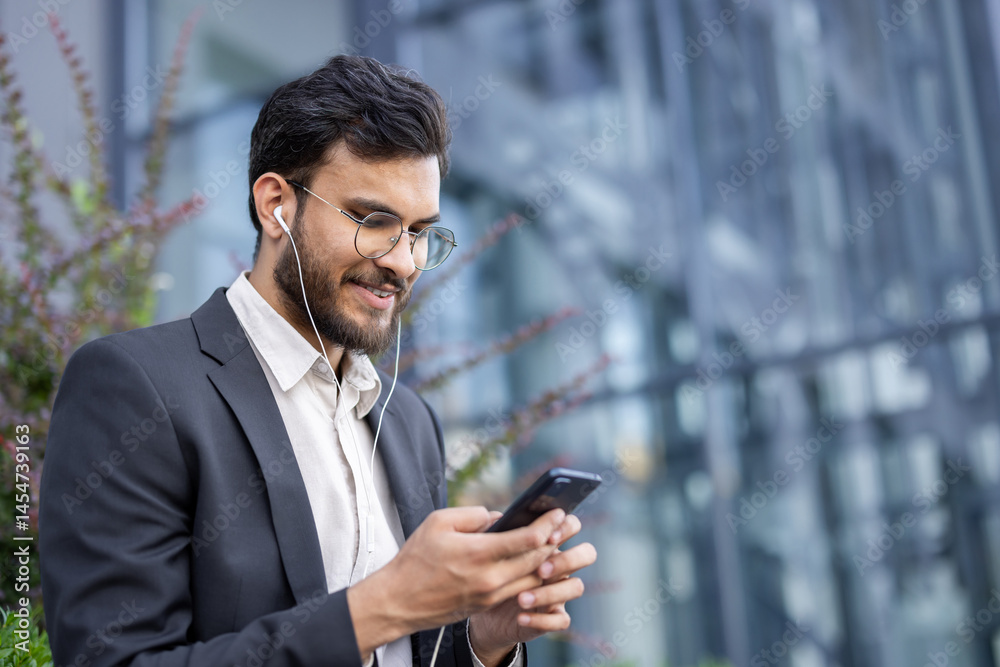 A young businessman in a suit smiles as he looks at his phone and listens with earphones near a modern building.