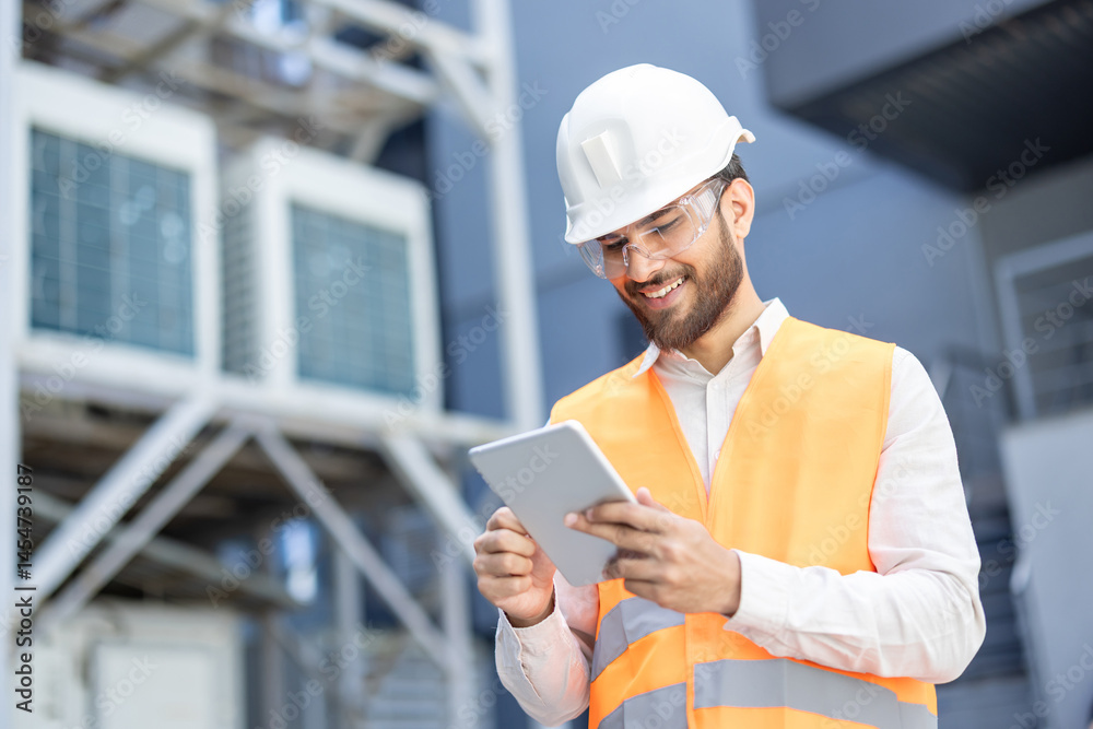 A smiling engineer in a white helmet and safety vest uses a tablet in a industrial setting, possibly for equipment inspections.