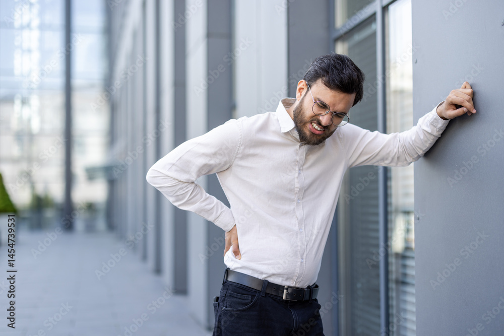 A businessman in a white shirt, glasses, and a beard is experiencing lower back pain outdoors near a building.