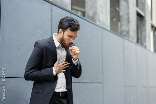 Obraz na płótnie A man in a suit is coughing, holding his chest, looking like he's experiencing discomfort or illness outdoors. w salonie