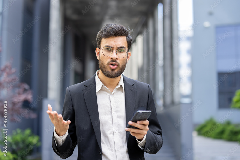A businessman, dressed in a suit, looks surprised and animated while holding a phone, gesturing with his hands in an outdoor setting.