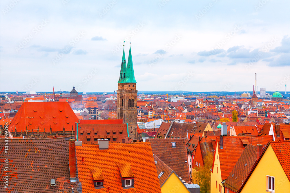 Panoramic view of Nuremberg showcases tiled rooftops, with twin green spires of Saint Lorenz Church rising above historic cityscape. Distant modern skyline blends with the historic architecture