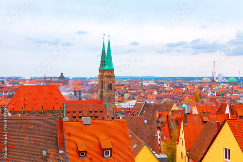 Panoramic view of Nuremberg showcases tiled rooftops, with twin green spires of Saint Lorenz Church rising above historic cityscape. Distant modern skyline blends with the historic architecture