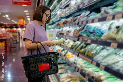 Woman choosing fresh vegetables in supermarket refrigerator shelf, holding shopping basket