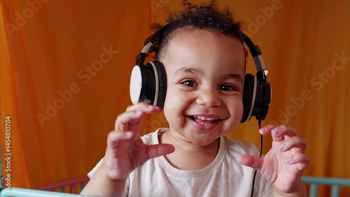 Adorable toddler wearing headphones and making heart gesture with hands, smiling in colorful nursery room