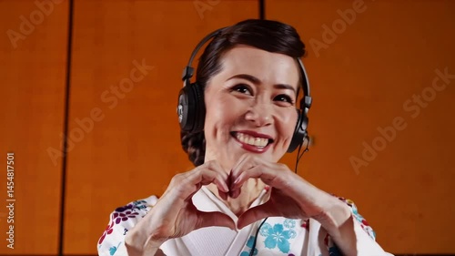 Smiling woman in traditional clothing with headphones making heart gesture with hands indoors