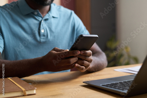 Medium closeup of unrecognizable African American man sitting at desk with laptop on it, scrolling social media page on smartphone