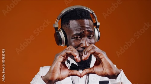 Emotional man in tuxedo with headphones making heart gesture with hands against orange background