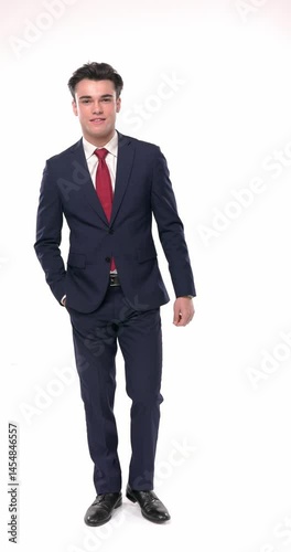 happy young businessman walking with hands in pockets and smiling while looking at camera in front of white background