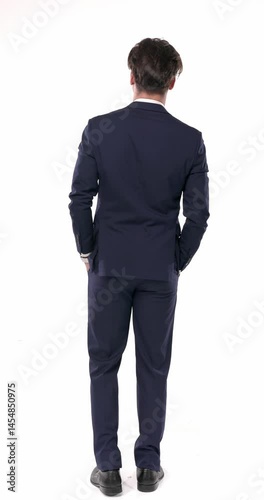 back view of elegant businessman in navy blue suit holding hands in pockets and moving in front of white background