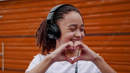 Smiling young woman with headphones making heart gesture with hands in front of orange garage door