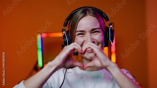 Smiling female gamer with headset making heart gesture with hands in front of colorful RGB-lit gaming setup