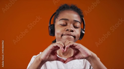 Young boy listening to music with headphones, forming a heart with hands against vibrant orange background