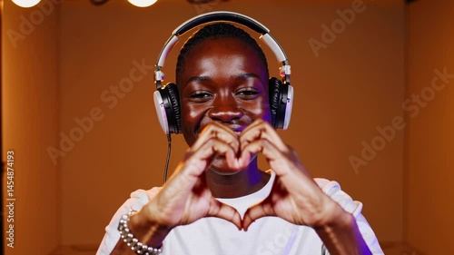 Smiling young man listening to music with headphones, making heart gesture with hands in warm indoor lighting