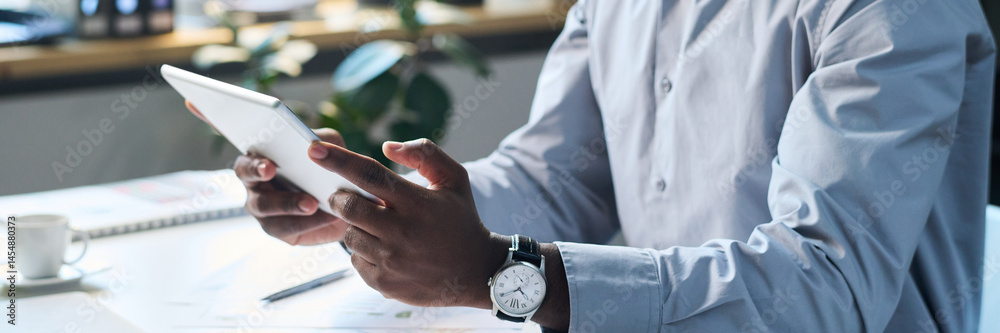 Medium closeup of modern black male manager sitting at desk in office, watching video on digital tablet, header