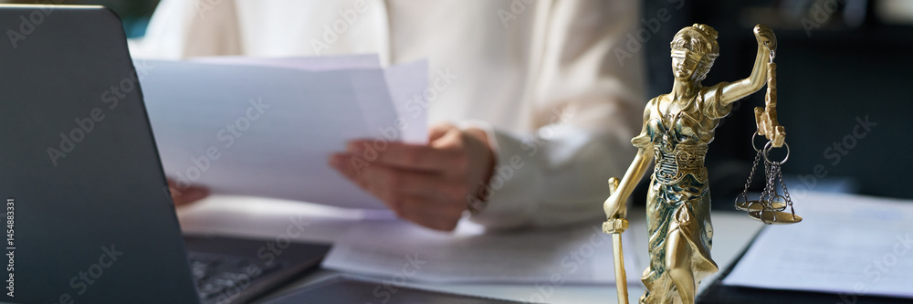 Bronze Lady Justice statuette on desk of attorney in law firm, holding scales and sword, and female lawyer working with documents in blurred background, header