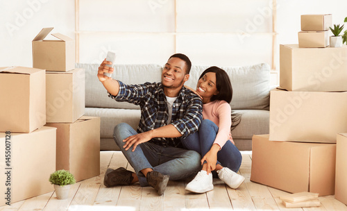 Smiling black guy and his girlfriend moving to their own home together, taking selfie among carton boxes, panorama. Happy African American couple making photo of first day in new apartment