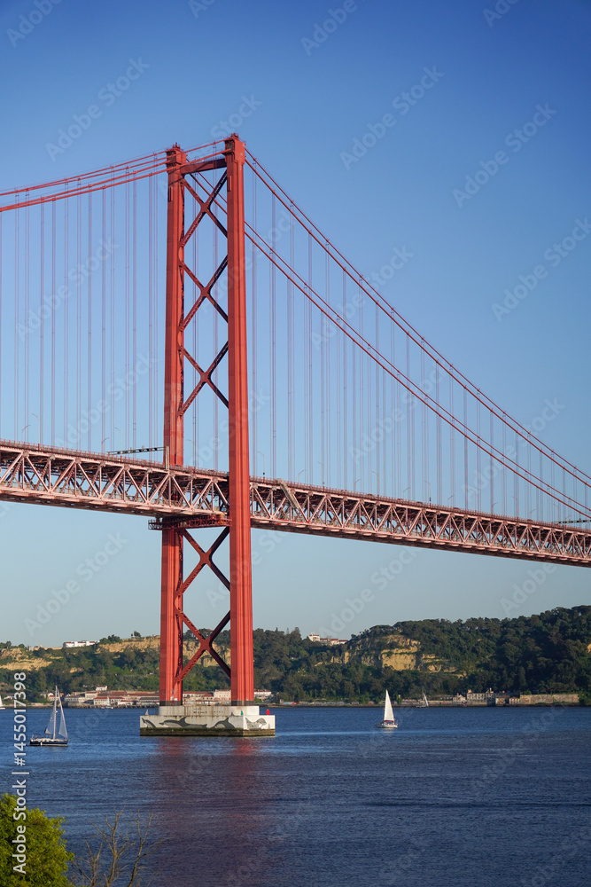 Fototapeta premium Majestic Red Suspension Bridge over Water with Sailboats Lisbon Portugal The Ponte 25 de Abril suspension bridge