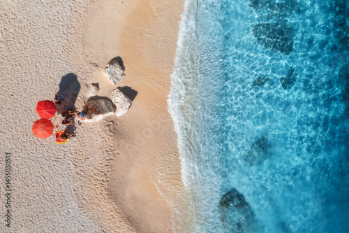 Fototapeta Naklejka Na Ścianę i Meble -  Aerial view of crystal-clear turquoise water gently laps the shore of a pristine sandy beach where tourists relax under red umbrellas. Picturesque summer scene. Top view of sandy beach, blue sea